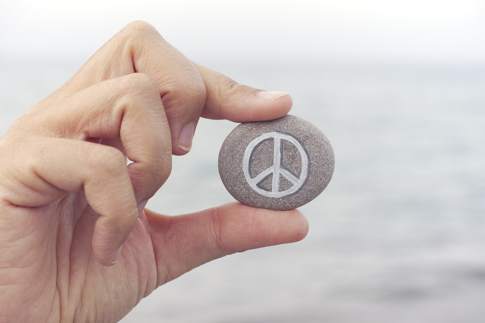 Person holds stone with peace symbol against sea in background. Close up.